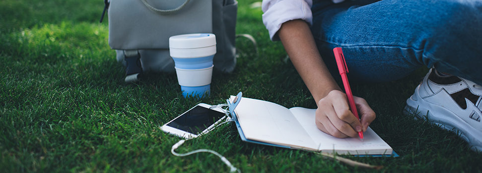A student with a backpack and coffee, writing notes in a notebook while sitting on grass.
